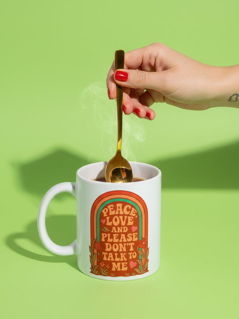A glossy white porcelain ceramic mug featuring a vibrant maximalist design centered on the exterior. The artwork displays a retro 1970s-inspired rainbow arch composed of concentric bands in burnt orange, mustard yellow, and teal green. Inside the arch, stylized vintage bubble-serif typography in reddish-orange reads PEACE LOVE AND PLEASE DON'T TALK TO ME. The design evokes a groovy, hippie aesthetic mixed with sassy introvert humor.
