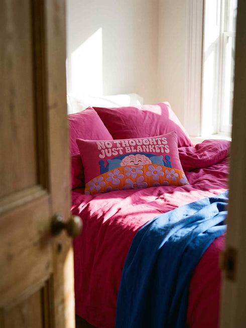 A bright magenta pink rectangular lumbar pillow cover featuring a flat 2D printed faux embroidery design. The printed illusion shows light pink bubble text reading NO THOUGHTS JUST BLANKETS. Below the text is a cartoon pink brain with a happy face, tucked into a blue bed with blue pillows. The brain is covered by a textured-looking orange blanket decorated with light purple daisies. The entire design is a smooth printed trompe l'oeil stitch effect, not real woven or stitched texture.