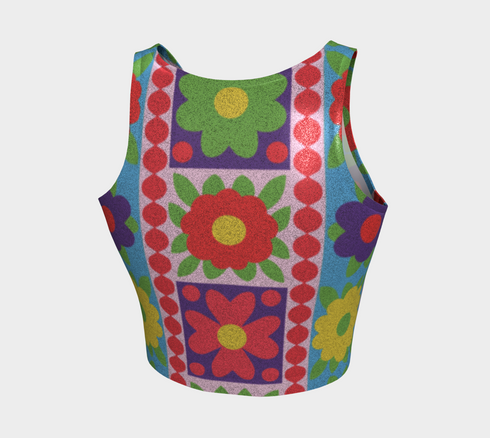 A high-resolution product photo of a body-hugging, 2D printed crop top with a 1970s-inspired floral patchwork pattern. The design consists of vibrant squares in cobalt blue, pale lavender, and deep violet, separated by pink borders with red dots. Central motifs include yellow daisies with orange centers and green-magenta floral mandalas. The top is made of a smooth, high-stretch performance knit fabric with curved back armholes and a cropped waistline.