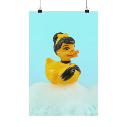 A chic rubber duck styled like Audrey Hepburn’s Holly Golightly, wearing a high bun, yellow bow, black gloves, and a sparkly choker. The duck floats in a pile of foam and is framed in white on a bubbly bathroom shelf with a red tile backdrop and soap dispenser.