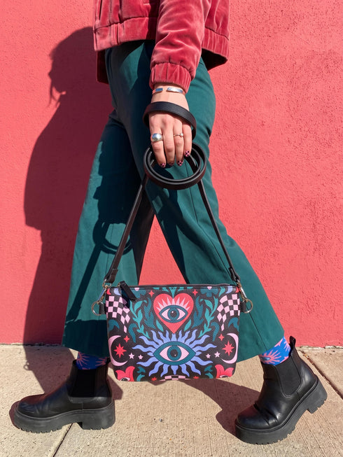 Small vegan leather crossbody purse with a real pebble grain texture and adjustable black strap. The flat printed artwork features a central blue sunburst evil eye motif below a coral pink sacred heart eye, surrounded by pink crescent moons, multicolored stars, flowing teal botanical vines, and wavy lavender and black checkerboard distortion banners. Set against a dark background with silver metal hardware including a top zipper, zipper pull, and strap clips. Measures 8.5 by 7 by 2 inches.