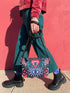 Small vegan leather crossbody purse with a real pebble grain texture and adjustable black strap. The flat printed artwork features a central blue sunburst evil eye motif below a coral pink sacred heart eye, surrounded by pink crescent moons, multicolored stars, flowing teal botanical vines, and wavy lavender and black checkerboard distortion banners. Set against a dark background with silver metal hardware including a top zipper, zipper pull, and strap clips. Measures 8.5 by 7 by 2 inches.