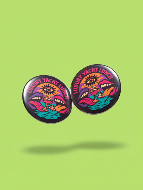 A round 1.75 inch glossy pinback button featuring satirical psychedelic artwork on a black background. The text LUXURY YACHT LUNCH arches over the top in bold neon pink letters. The central design depicts a surreal scene with a stylized third-eye sun, large open mouths with tongues sticking out, a smiling cocktail glass character, and a small white yacht floating on wavy teal water. The color palette includes retro 70s neon pink, orange, purple, and teal. The button has a safety pin backing.