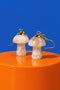 Two white mushroom-shaped earrings with gold accents are displayed on an orange surface against a blue background.