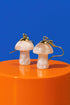 Two white mushroom-shaped earrings with gold accents are displayed on an orange surface against a blue background.