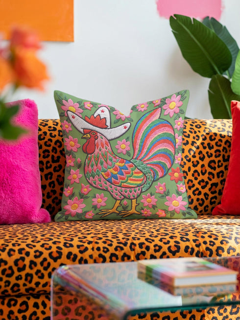 Square throw pillow cover with a printed faux embroidery design of a rooster wearing a white cowboy hat with red stars on a grass green background. The western chicken has a red comb and wattle, yellow beak and feet, and layered feathers in pink, green, orange, blue, and yellow with a rainbow striped tail. Pink and red flowers with yellow centers frame the bird. The stitched look is a flat 2D print on a smooth linen feel polyester cover with a hidden zipper, not raised embroidery.