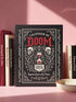 An 8.5 x 11 inch black hardcover spiral planner with a durable satin matte laminated finish. The front cover features a vintage-style white ornamental border, creating a gothic, macabre aesthetic. Inside the frame, the title "Calendar of Doom" is printed in an antique serif font. This undated weekly and monthly agenda is shown with a metal wire-o spiral binding on the right side. The design uses a black, white, and red color scheme, making it a unique piece of dark humor stationery