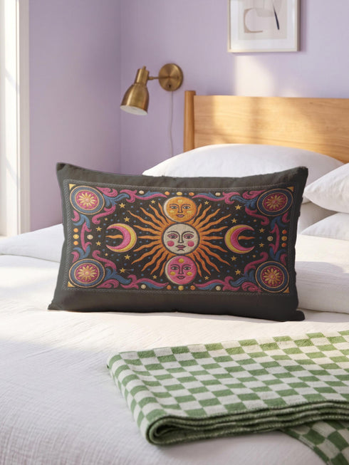 Rectangular 20x12 inch lumbar pillow cover in black featuring printed faux crewel embroidery of a large orange sunburst with a cream face, flanked by pink and yellow crescent moons, a golden moon face above and a pink moon face below, gold stars, blue dots, magenta and blue baroque scroll flourishes, and four corner medallions with golden flowers. Whimsigoth celestial design printed on both sides with a hidden zipper, crafted from soft linen look polyester.
