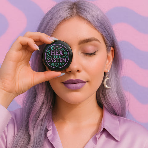Glossy black pinback button with pastel embroidery-style design that says “Hex the System,” surrounded by vines, stars, and a crescent moon. Displayed against witchy pink and blue backdrops, street pole flyers, and protest props.