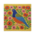 Square faux embroidery throw pillow cover with a large stylized folk art bird facing left. The bird has a cobalt blue head, orange belly, light blue tail, and pink legs, set on a bright mustard yellow background scattered with pink and red daisy flowers on trailing green vines. The smooth, linen-feel polyester uses a printed trompe loeil effect to mimic raised thread needlework. Bold maximalist folk art bird pillowcase and cottagecore cushion cover for eclectic, colorful, dopamine decor homes.