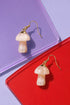 two pairs of mushroom-shaped earrings, one in gold and the other in white, placed on a red and purple background.