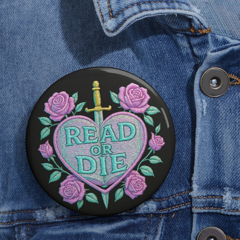 Glossy black pinback button featuring embroidered-style artwork of a pastel purple heart that reads “Read or Die” pierced by a sword and surrounded by roses; shown on books, graffiti, and lace backgrounds.