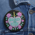 Glossy black pinback button featuring embroidered-style artwork of a pastel purple heart that reads “Read or Die” pierced by a sword and surrounded by roses; shown on books, graffiti, and lace backgrounds.