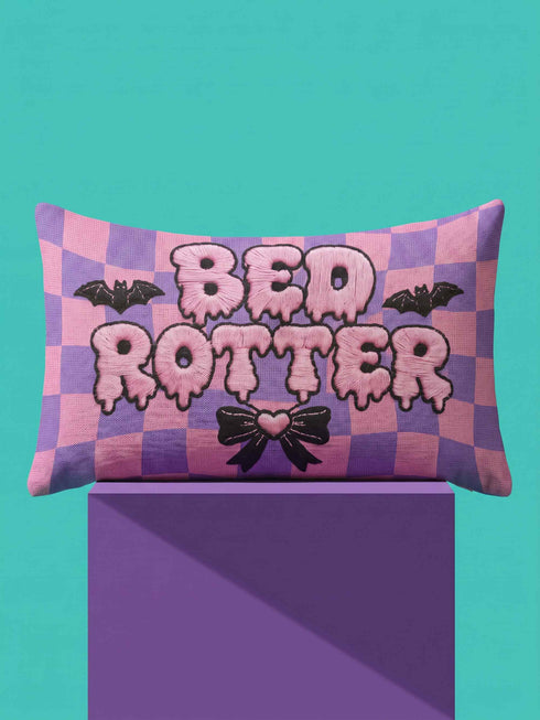 Pastel goth lumbar pillow, 20 by 12 inches, with a pink and lavender purple wavy checkerboard background. The words Bed Rotter appear in dripping pink bubble letters with a faux embroidery thread texture and black outlines. Two black bats with pink stitch details flank the text, and a black bow with a pink heart center sits below. Double sided flat print that mimics raised embroidery on linen feel polyester with a hidden zipper, insert included.