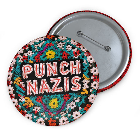 Round pinback metal button with an embroidered-style design on a black background. The center displays the phrase "PUNCH NAZIS" in bold, white stitched-textured lettering with red accents. Surrounding the text is an elaborate folk-inspired floral pattern, densely packed with embroidered-style flowers in shades of red, yellow, blue, pink, and white. The intricate design includes symmetrical lace-like stitching and decorative motifs, creating a rich, tapestry-like effect. The button has a silver metal back wi