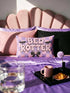 Pastel goth lumbar pillow, 20 by 12 inches, with a pink and lavender purple wavy checkerboard background. The words Bed Rotter appear in dripping pink bubble letters with a faux embroidery thread texture and black outlines. Two black bats with pink stitch details flank the text, and a black bow with a pink heart center sits below. Double sided flat print that mimics raised embroidery on linen feel polyester with a hidden zipper, insert included.