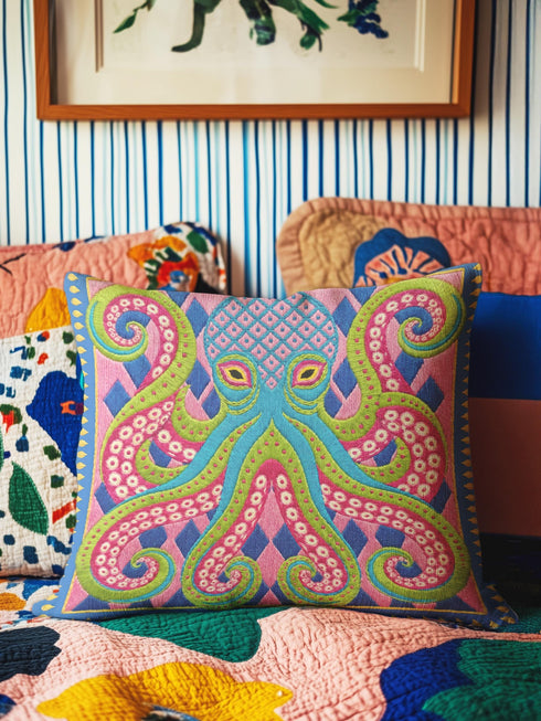 A square decorative throw pillow cover featuring a highly symmetrical bright octopus design. The octopus has a turquoise blue body with a diamond patterned head and eight curled tentacles detailed in bright pink and lime green. The background is a repeating diamond argyle pattern in royal blue and light pink. The artwork is a flat two dimensional print that mimics the textured look of intricate crewelwork embroidery. The pillow features a hidden zipper on the bottom edge. No insert included.