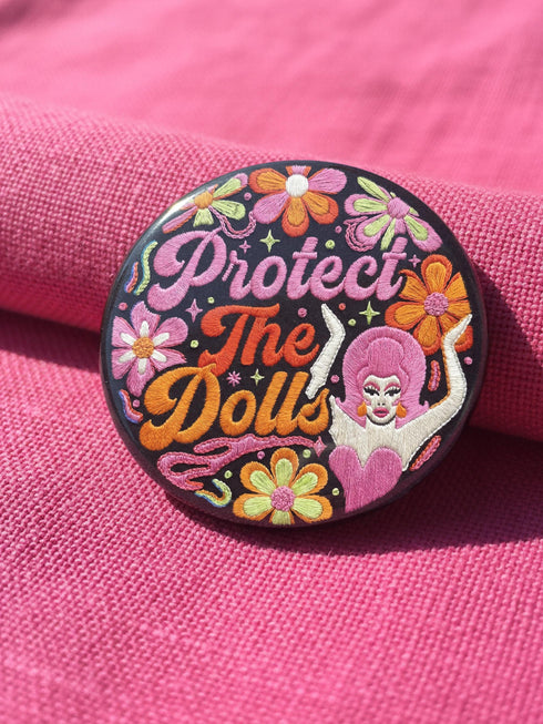 A circular black pinback button featuring the slogan Protect The Dolls in retro script. The design utilizes a high-fidelity trompe-l'œil print style that mimics the texture of raised thread embroidery with satin-stitch detailing. The artwork features vibrant hot pink, rust orange, and golden yellow motifs including drag queens with bouffant hair, 70s style daisies, lightning bolts, and a crescent moon with a face. The surface is smooth and glossy, creating a faux-textured illusion.