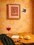A vibrant fine art print of a rubber duck inspired by Frida Kahlo, complete with bold unibrow, floral crown, and traditional embroidered blouse. Framed in a red sculptural frame, the duck floats in orange-tinted bubbles against a deep orange backdrop and violet wall.