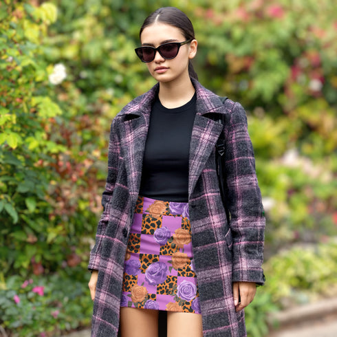 A woman wearing a purple plaid coat, sunglasses, and a patterned skirt stands in a garden surrounded by greenery.