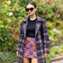 A woman wearing a purple plaid coat, sunglasses, and a patterned skirt stands in a garden surrounded by greenery.