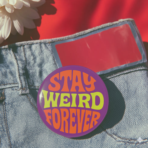 A round metal pin button with a glossy finish and a vibrant purple background. The phrase 'STAY WEIRD FOREVER' is written across the center in a wavy, psychedelic 70s-style font. The words 'STAY' and 'FOREVER' are a bold, bright orange, while the word 'WEIRD' is rendered in a contrasting, electric chartreuse lime green. The button has a smooth, scratch-resistant mylar surface covering the design. The pin is shown in its 2-inch and 3-inch variations, highlighting its circular shape and durable construction.