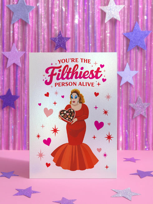 A 5x7 inch pearlescent silver greeting card featuring a flat vector illustration of the drag queen Divine in a red mermaid gown holding a heart-shaped box of chocolates. The text above reads "You're The Filthiest Person Alive" in bold red script with pink sparkles. The card has a shimmering finish that reflects light. Inside text reads "...and I love it." The back features the Honey Dazed logo. Includes a matching envelope.