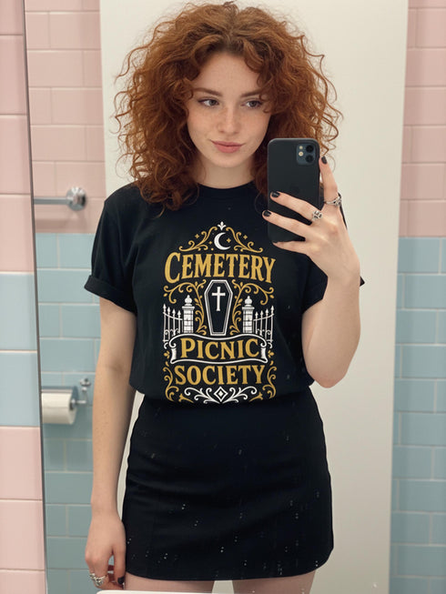 A black unisex crewneck graphic t-shirt featuring an ornate Victorian-inspired emblem printed on the chest in vintage gold and off-white. The centralized, symmetrical design showcases a classic coffin with a cross on the lid, flanked by wrought-iron cemetery gates. Beautiful scrollwork and decorative filigree flourishes surround the bold, retro serif text reading CEMETERY PICNIC SOCIETY. A crescent moon and stars sit at the top, completing this gothic memento mori graphic shirt.