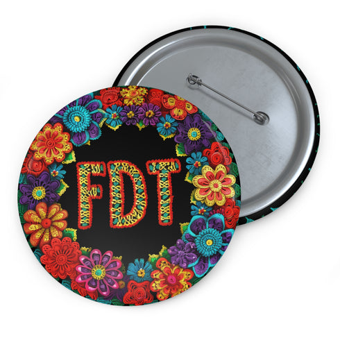 Circular black pinback button with bright 'FDT' lettering in orange/teal surrounded by colorful embroidery-style flowers in red, purple, blue, and yellow. Metal safety pin backing visible on reverse side. Political protest pin with vibrant Mexican folk art-inspired floral design.