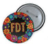 Circular black pinback button with bright 'FDT' lettering in orange/teal surrounded by colorful embroidery-style flowers in red, purple, blue, and yellow. Metal safety pin backing visible on reverse side. Political protest pin with vibrant Mexican folk art-inspired floral design.