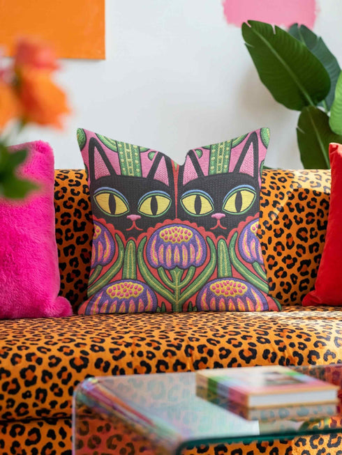 A square decorative pillow cover featuring a vibrant, symmetrical flat printed design that mimics the look of textured embroidery. Two black cats with large yellow eyes and pink noses face forward against a bright red background. Below the cats are large purple and pink flowers with green stems. Above the cats are green cactus shapes. The artwork uses bold, colorful outlines to create an optical illusion of stitching, though the fabric itself is smooth.