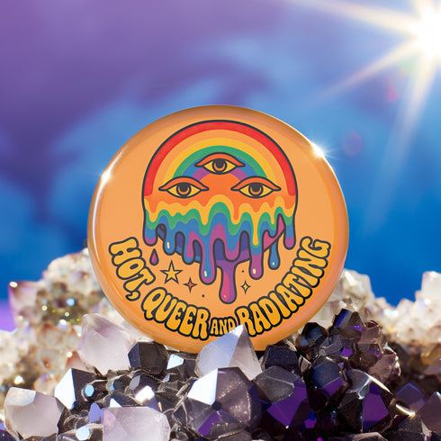 Round orange pinback button featuring bold rainbow psychedelic artwork of a melting third-eye face beneath a rainbow arch. Surrounded by stars and the phrase “Hot, Queer and Radiating” in retro black bubble letters. Glossy metal badge with UV-resistant mylar face and secure safety pin back.