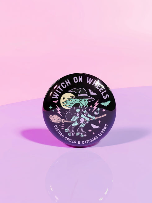 A round pin with a cartoon illustration of a witch riding a broomstick, surrounded by various Halloween-themed elements such as bats, skulls, and a crescent moon.