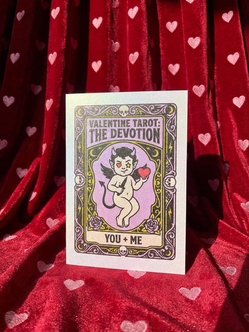 A vertical folded greeting card featuring a pastel goth tarot design titled The Devotion. The central illustration shows a pale devil imp with black wings and horns, holding a red heart in the left hand and a black dagger in the right. The imp has one star eye and one heart eye. The background is lavender purple. A black border frames the scene with teal vines, skulls, gold crescent moons, and roses. The bottom banner reads YOU + ME. The finish is shimmering pearlescent.