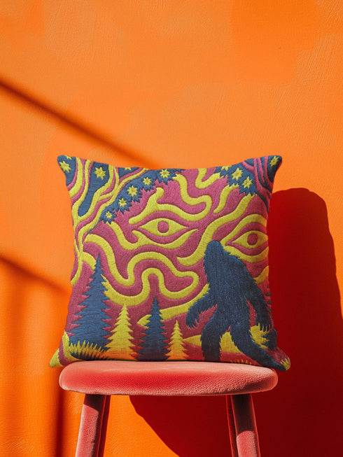 A square decorative throw pillow cover featuring a trippy retro Bigfoot scene. A large navy blue silhouette of Sasquatch walks through a forest of navy and yellow pine trees. The background is a psychedelic swirling sky in vibrant pink and yellow, featuring a large stylized eye and a cluster of small yellow stars. The flat two dimensional print visually mimics the textured look of heavy needlepoint embroidery. The pillow features a sleek hidden zipper on the bottom. No insert is included.