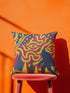 A square decorative throw pillow cover featuring a trippy retro Bigfoot scene. A large navy blue silhouette of Sasquatch walks through a forest of navy and yellow pine trees. The background is a psychedelic swirling sky in vibrant pink and yellow, featuring a large stylized eye and a cluster of small yellow stars. The flat two dimensional print visually mimics the textured look of heavy needlepoint embroidery. The pillow features a sleek hidden zipper on the bottom. No insert is included.