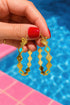 A hand holding a pair of gold hoop earrings with diamond-shaped designs and turquoise accents, with a blurred background of a swimming pool.