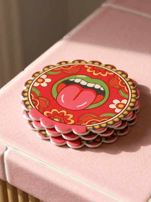 Round ceramic coaster with scalloped edges featuring a vibrant pop art design of an open mouth with green lips and a red tongue sticking out. The background is a bright red with psychedelic floral swirls in yellow orange and green. The surface is glossy glazed ceramic. The back is made of cork to prevent slipping. This maximalist decor piece fits retro 70s funky and weirdcore aesthetics. It is a bold statement piece for coffee tables. The design is colorful and eclectic dopamine decor.