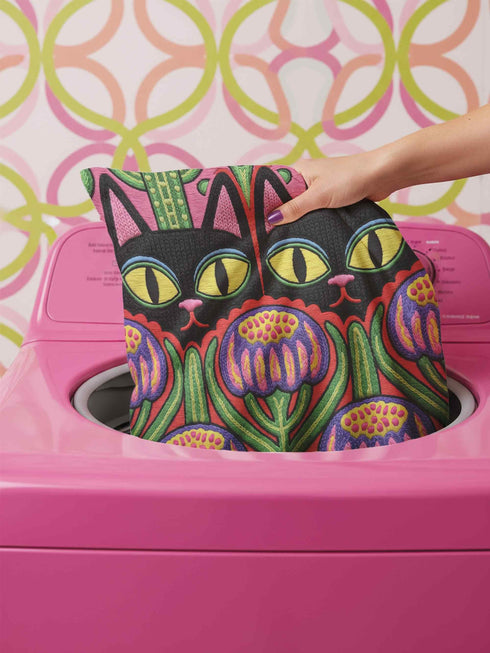 A square decorative pillow cover featuring a vibrant, symmetrical flat printed design that mimics the look of textured embroidery. Two black cats with large yellow eyes and pink noses face forward against a bright red background. Below the cats are large purple and pink flowers with green stems. Above the cats are green cactus shapes. The artwork uses bold, colorful outlines to create an optical illusion of stitching, though the fabric itself is smooth.