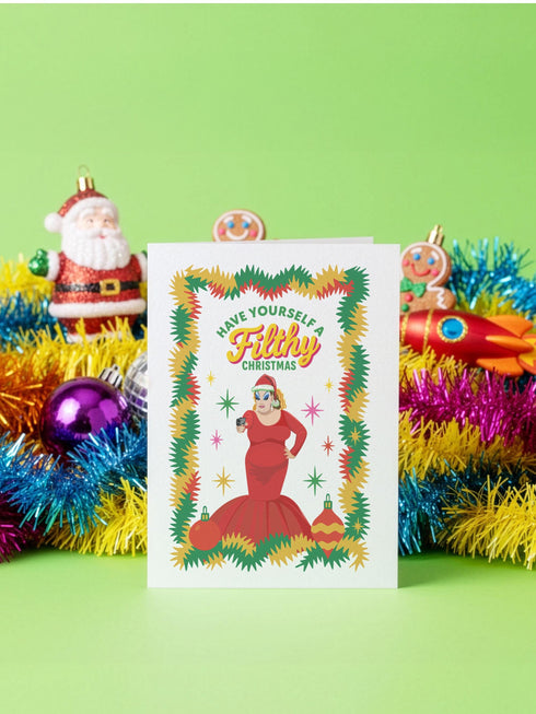 A 5x7 pearlescent holiday card by HoneyDazed. The card illustration, inspired by John Waters, features the drag queen Divine in a red dress with a Santa hat. She points a gun, withtext reads "Have Yourself a Filthy Christmas" in campy font. The card is bordered by garland. Iinside of the card reads: "Season's Sleazings" in red and green text. This kitschy greeting card is a perfect piece of cult film memorabilia and a funny adult humor gift for fans of queer art.