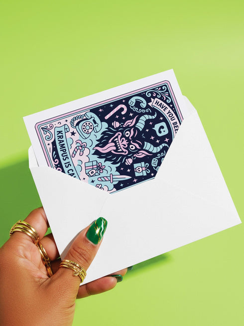 A vertical 5x7 holiday card featuring a retro tattoo style illustration of Krampus in pastel pink, teal, and black ink on shimmery silver pearlescent paper. The central horned demon has a long tongue and is surrounded by vintage rotary phones, daggers, candy canes, and skull gifts. Top text asks Have You Been Good while the bottom banner reads Krampus Is Calling. The interior text reads Let's be honest... Being good is overrated. Merry Christmas.