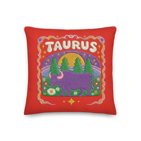 A square vibrant red throw pillow featuring a central illustration of the Taurus zodiac sign. The word TAURUS appears at the top in white bubbly letters with a faux-stitch texture. Below the text is an arch containing a purple bull standing in a green field dotted with white daisies and red flowers. Behind the bull are green pine trees and a large yellow sun against a blue sky. The design is framed by a pink wavy border, printed to look like raised embroidery on linen-textured polyester fabric.