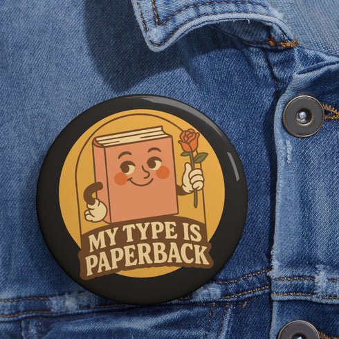 A glossy black round pin-back button with a retro 1970s-style design. The center shows a smiling cartoon paperback book holding a red rose, colored light brown with white gloves and feet. Below, “MY TYPE IS PAPERBACK” appears in chunky brown-and-tan 70s lettering. This bookish flair piece has a high-gloss, scratch-resistant mylar finish, secure safety pin backing, and comes in two- or three-inch sizes.