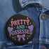 Glossy black pinback button featuring pastel-style text that reads “Pretty and Possessed” with a bow, candles, hearts, and sparkles.