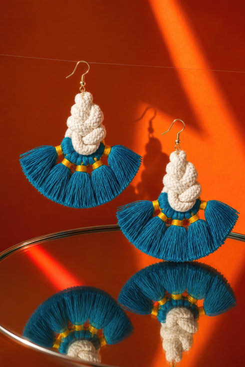 a pair of earrings with blue and white tassels, displayed on a reflective surface against an orange background.