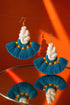 a pair of earrings with blue and white tassels, displayed on a reflective surface against an orange background.
