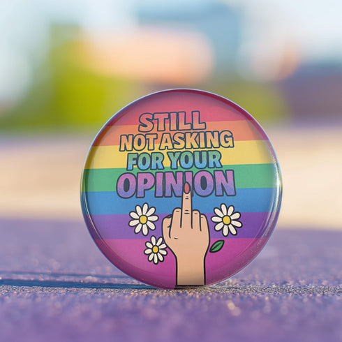 Round glossy pinback button measuring 2.25 inches with a bold rainbow pride flag background. Centered text reads “Still Not Asking For Your Opinion” in black, yellow, blue, and purple letters. Below the text is an illustrated hand with painted red nails giving the middle finger, surrounded by white daisies. The design is printed on tinplate with a scratch- and UV-resistant mylar coating. Button features a strong safety pin backing, perfect for bags, jackets, or pride outfits.