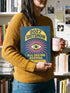 AA hardcover spiral planner for 2027 with a dark blue cover and retro 70s style graphics. The title reads "2027 SHITSHOW All-Seeing Agenda" in bold yellow and pink text. Below the title is a large, stylized all-seeing eye with pink rays emanating from it. At the bottom, a pink ribbon contains the text "BLINK TWICE FOR MERCY." The back cover is also dark blue with a yellow sun graphic and the text "Plans bloom. Deadlines hum. I write, I wander, then get it done. Keep receipts. Keep grace.