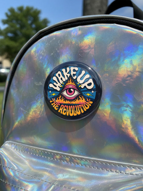 A round pinback button featuring a retro psychedelic design. The central focus is a stylized Eye of Providence in a pink triangle, surrounded by vibrant orange and yellow flames. Behind the triangle are teal and petrol blue sunburst rays. The text "WAKE UP" curves across the top in cream wavy letters, and "THE REVOLUTION" curves along the bottom in orange. The button has a glossy finish and is available in 1.75 inch or 2.25 inch sizes.