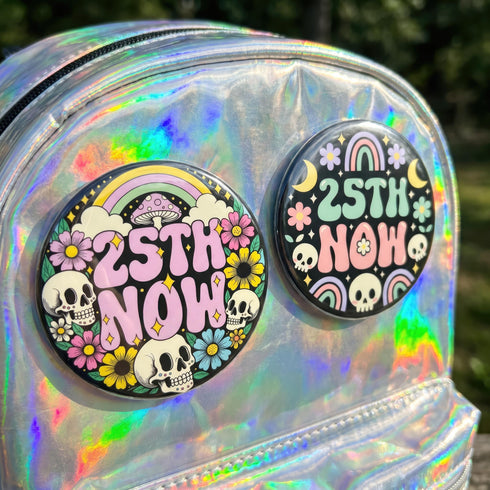 A round pinback button featuring a solid black background covered in a retro pastel colored design. The center reads 25TH NOW in a groovy bubbly font using pale pink and mint green referencing the 25th amendment. Surrounding the text is a maximalist cluster of cute but edgy motifs including smiling white skulls, pastel rainbows, pale yellow crescent moons, daisies, and a pink and white spotted mushroom. The button has a glossy laminate finish and a secure metal safety pin backing.